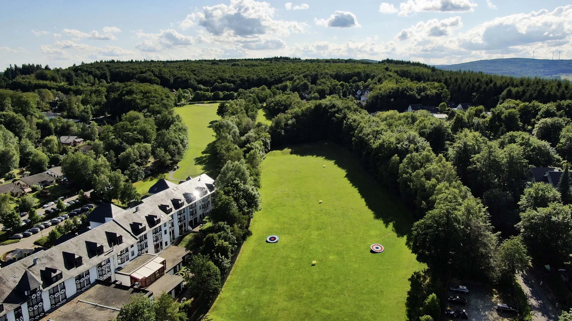 Luftaufnahme eines grünen Bogensportplatzes neben einem Hotel umgeben von Wald