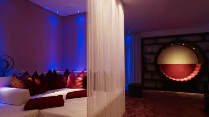 Lounge mit weißen Sitzmöbeln, rotem Kissen, Raumteiler und runder Sitzecke bei blauem Licht