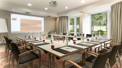 Konferenzraum mit U-förmigen Tischen, Stühlen, Wasserflaschen und roten Äpfeln