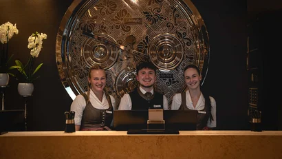 Drei lächelnde Hotelmitarbeiter am Empfangstresen mit dekorativer Uhr im Hintergrund